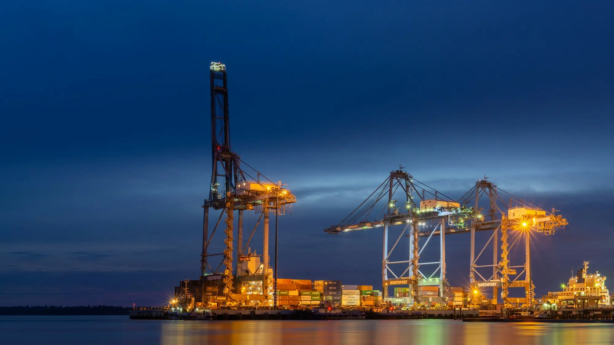 Container cranes lit up at dusk over stacked shipping containers at a container terminal, where imported stock waits for customs clearance
